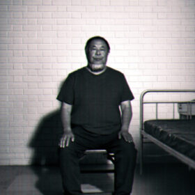 A black-and-white image of Ai Weiwei sitting upright on a small stool in a sparse, cell-like room. He faces the camera with his hands resting on his knees. Behind him is a white brick wall, and to his right there is a simple metal bed frame with a thin mattress. The lighting is stark, casting strong shadows on the wall and floor, giving the scene a stark, surveillance-like atmosphere