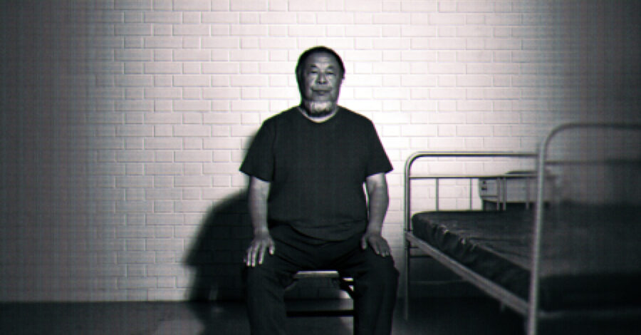 A black-and-white image of Ai Weiwei sitting upright on a small stool in a sparse, cell-like room. He faces the camera with his hands resting on his knees. Behind him is a white brick wall, and to his right there is a simple metal bed frame with a thin mattress. The lighting is stark, casting strong shadows on the wall and floor, giving the scene a stark, surveillance-like atmosphere