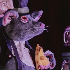 A performer dressed up as a mouse, wearing clothes and holding a wedge of cheese.