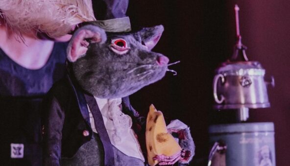 A performer dressed up as a mouse, wearing clothes and holding a wedge of cheese.
