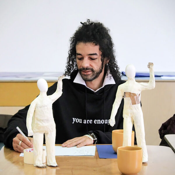 A man sits at a table sketching. There are two ceramic figures on the table top.