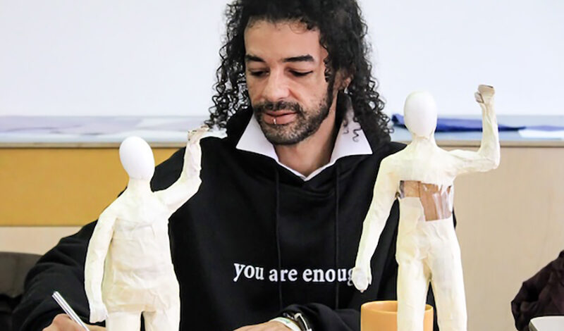 A man sits at a table sketching. There are two ceramic figures on the table top.
