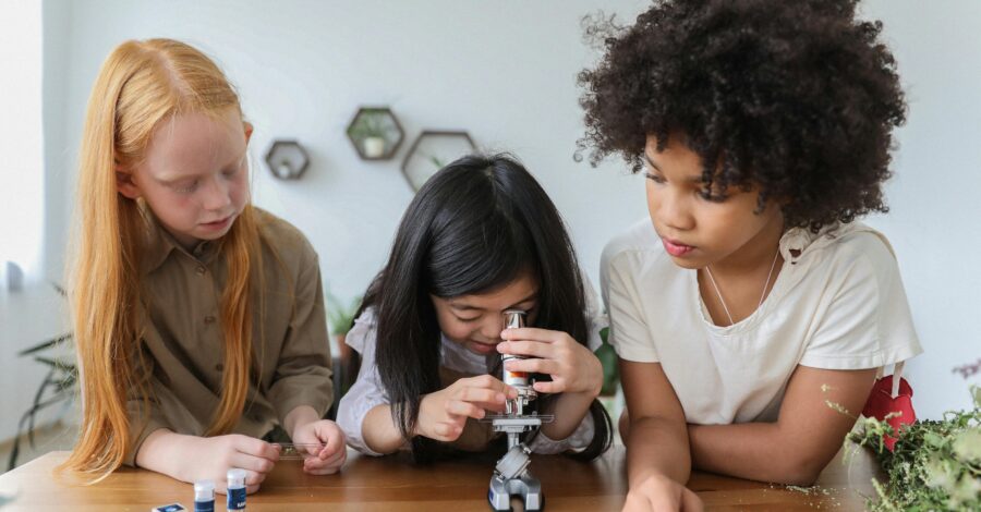 A little girls looks through a microscope while two other girls look on.
