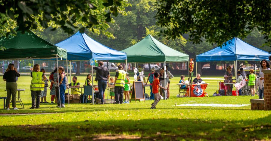 A row of different coloured gazebos in a park and there are people standing around them.