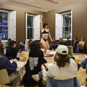 A woman performs in the middle of the room. The audience are sat on chairs around her. She is wearing a black bikini.