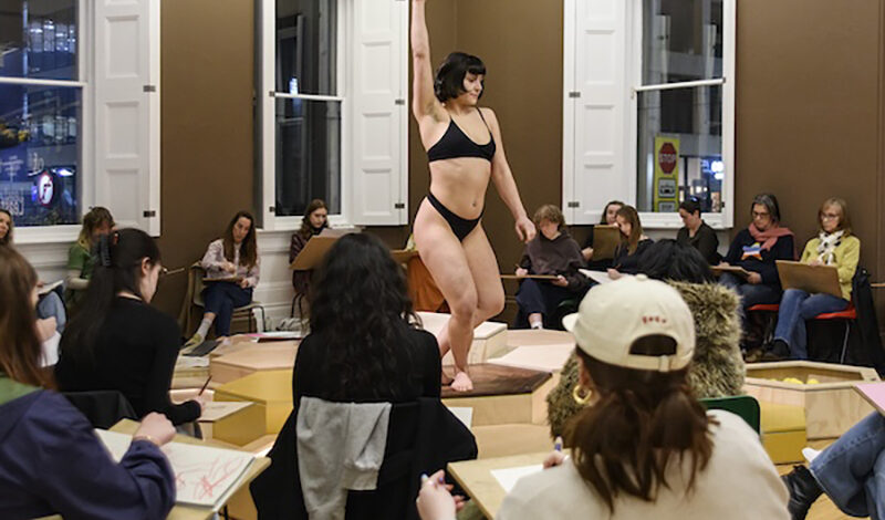 A woman performs in the middle of the room. The audience are sat on chairs around her. She is wearing a black bikini.