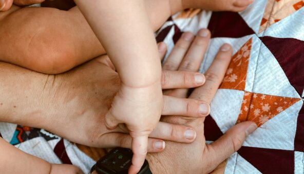 A close-up of a baby's chubby hand resting on stacked adult hands, with a tiny finger gently touching the screen of a black smartwatch.