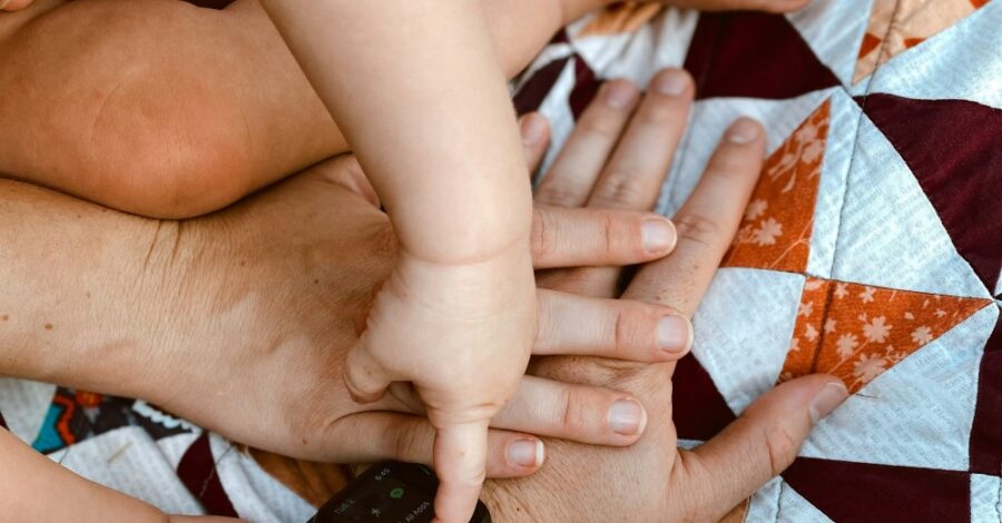 A close-up of a baby's chubby hand resting on stacked adult hands, with a tiny finger gently touching the screen of a black smartwatch.