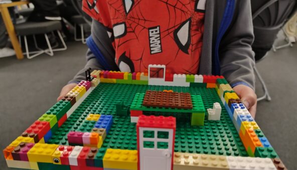 A child wearing a spider man t shirt holds up a Lego house.