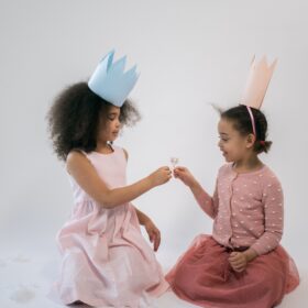 Two little girls wearing princess paper hats exchange flowers.