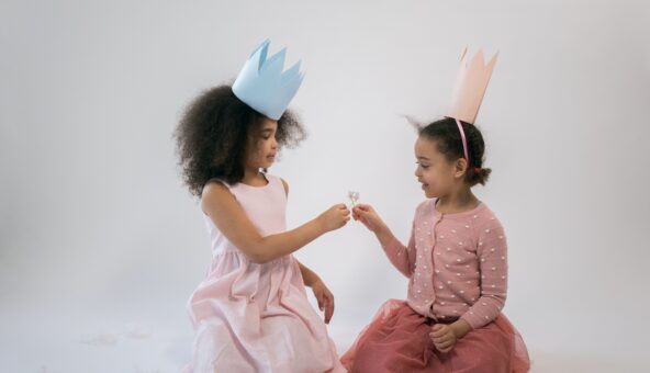 Two little girls wearing princess paper hats exchange flowers.