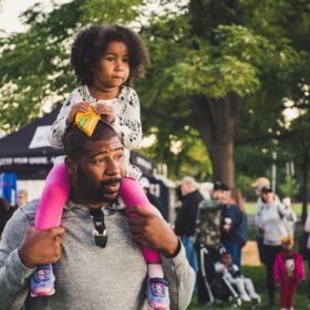 A man carrying a child on his shoulders at an event in a park.