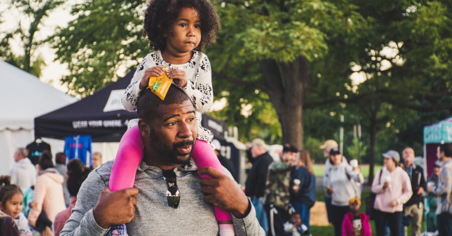 A man carrying a child on his shoulders at an event in a park.