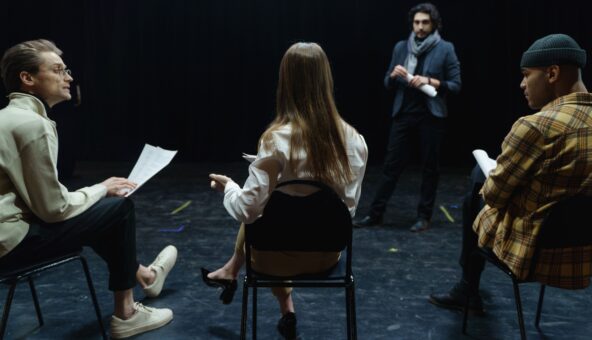 Three people sat on chairs in a black box studio. One person is stood up as if they are teaching the class.