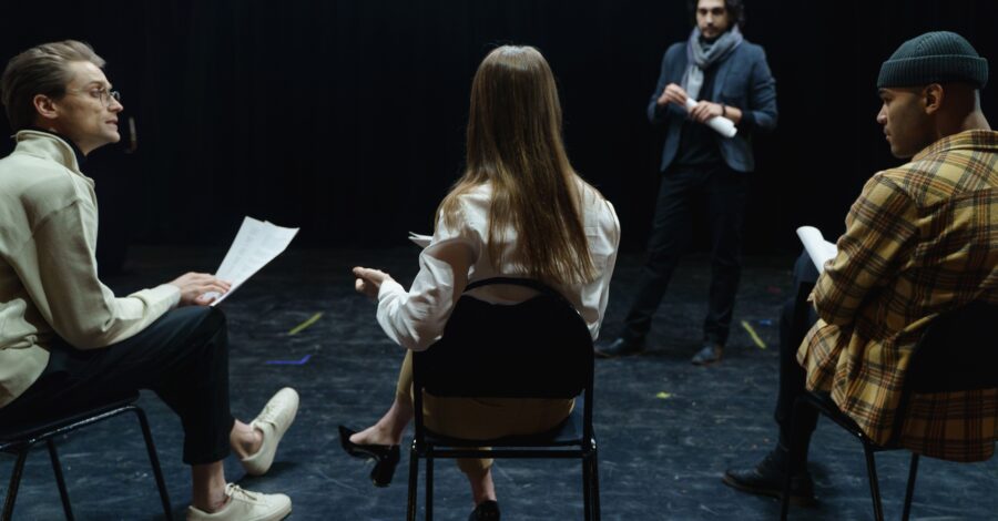 Three people sat on chairs in a black box studio. One person is stood up as if they are teaching the class.