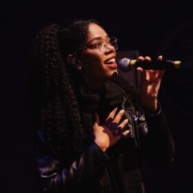A woman with long black hair, standing, speaking into a microphone.