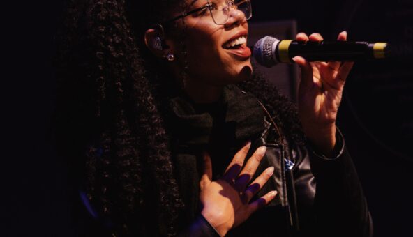 A woman with long black hair, standing, speaking into a microphone.