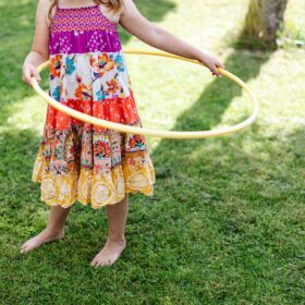 A girl in a red and white floral dress standing on green grass holding a yellow hula hoop.
