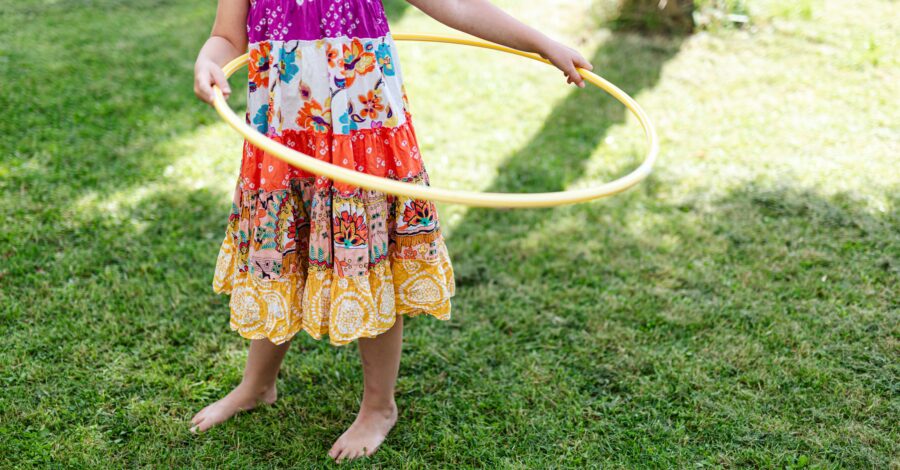 A girl in a red and white floral dress standing on green grass holding a yellow hula hoop.