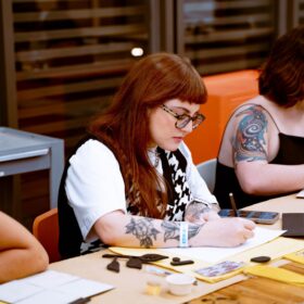 Three people sat at a table writing on a pad with a pen.