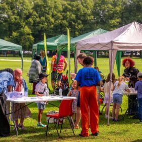 People are stood under colourful gazebos on a sunny day in a Manchester park.