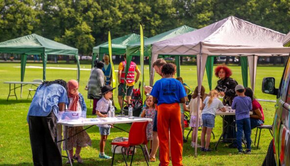 People are stood under colourful gazebos on a sunny day in a Manchester park.