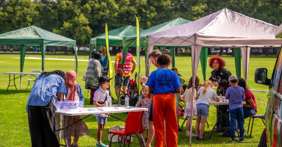 People are stood under colourful gazebos on a sunny day in a Manchester park.